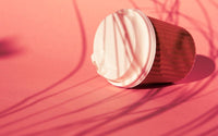 Mug en carton avec couvercle en plastique blanc renversé sur un fond rose, avec des ombre d'herbes hautes.