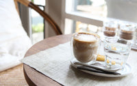 Tasse à café à double paroi posée sur une assiette avec une serviette papier et un verre à eau. Sur une table ronde en bois avec un set de table.