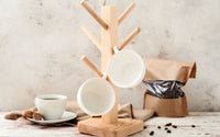 Support porte tasses à café en bois vertical. Deux tasses y sont accrochées. En arrière plan un sac de café en grain à droite, et une tasse et sous-tasse blanche à gauche.