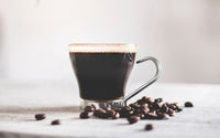 Tasse à café en verre avec une poignet métal, remplie de café et posée sur une table blanche à côté de grains de cafés.