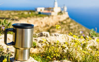 Tasse thermos gris métal avec la poignet et le couvercle noir, posée sur un rocher, avec une vue floutée sur un château en contre-bas 