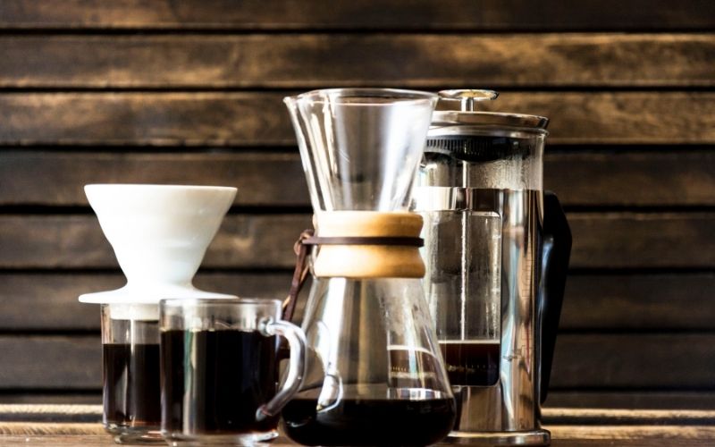 une cafetière à piston en verre, une cafetière à extraction lente en verre et deux tasses en verre dont une avec un filtre réutilisable en porcelaine dessus.