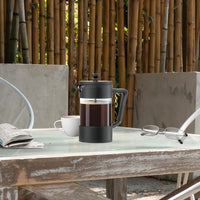 Cafetière Italienne à Piston Résistante et Facile d'Utilisation sur une tableau en extérieur avec tasse de café et lunette à côté et bambou en arrière plan
