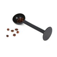Cuillère Doseuse Noire avec Presse Poudre, sur un fond blanc, avec des graines de café.