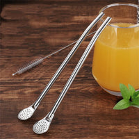 Cuillère à Thé Polyvalente Argenté en Forme de Paille posée contre un verre de jus d'orange et sur une table en bois