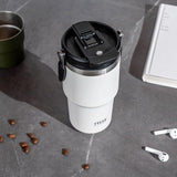 Gourde Isotherme 750ml Forme Tasse, posée sur un bureau gris foncé à côté 'un livre, une paire d'écouteur sans fil et des grains de café.