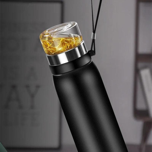 Gourde Isotherme 750ml avec Infuseur à Thé, tenue par sa sangle, on voit dans la partie en verre le thé avec de l'eau qui à colorée.